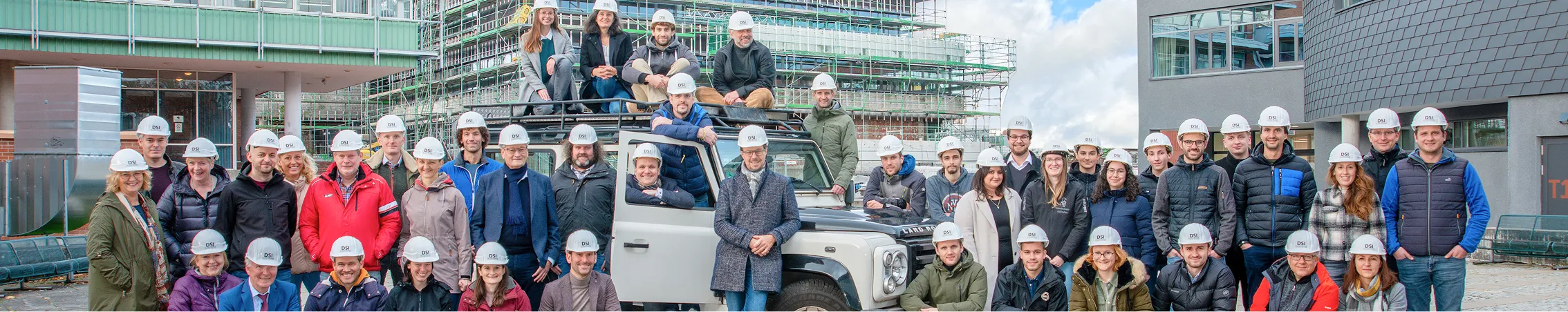 Große Gruppe von Menschen mit weißen Schutzhelmen steht vor einem weißen Geländewagen und einem im Bau befindlichen Gebäude.