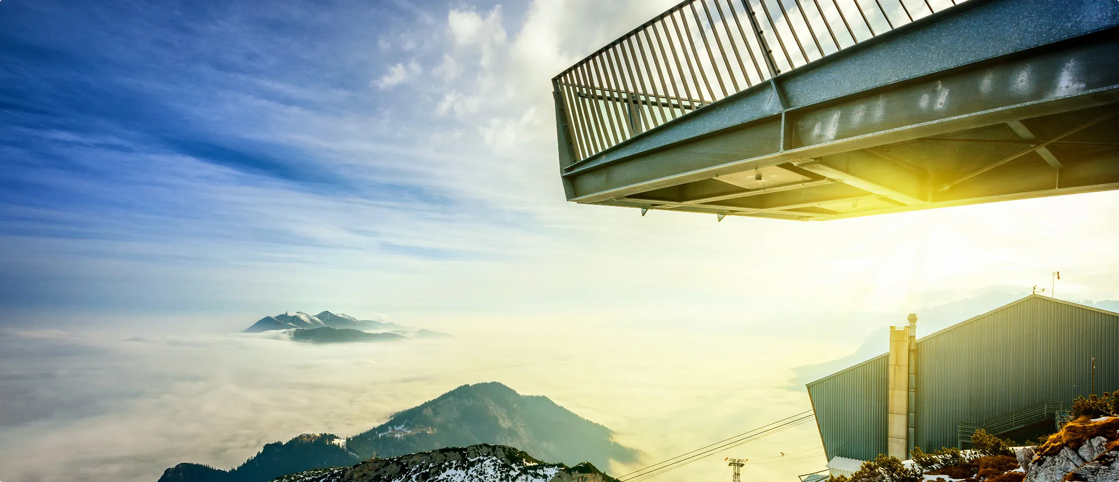 Berggipfel über einer Wolkendecke bei Sonnenaufgang mit einem Stahlbalkon und einem Gebäude im Vordergrund.