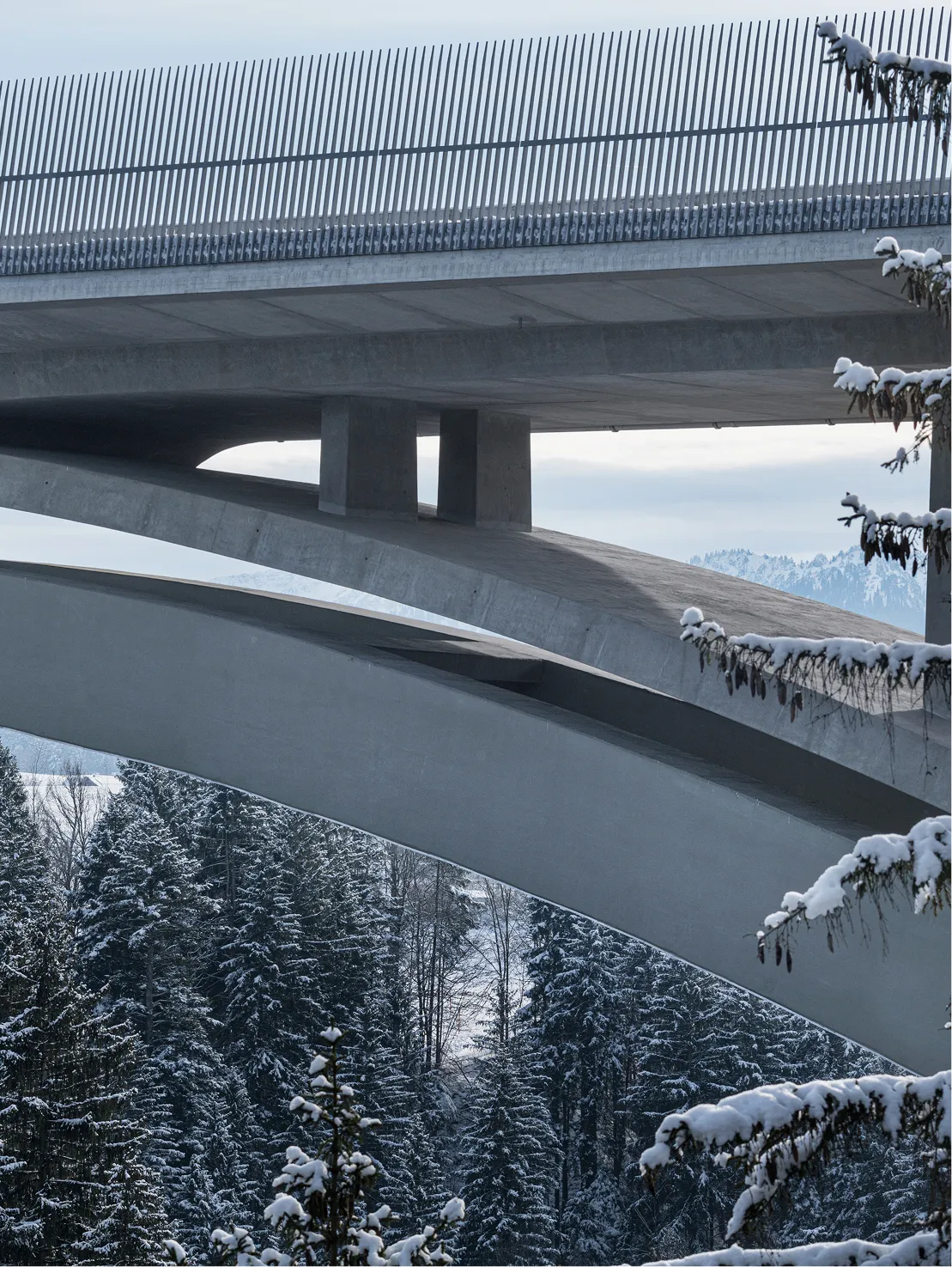 Nahaufnahme einer modernen Betonbrücke mit schneebedeckten Tannenbäumen im Winter unter ihr.