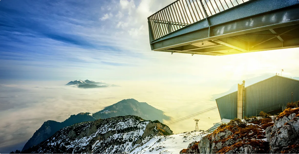 Berglandschaft mit schneebedeckten Gipfeln, einer Seilbahnstation und einem Aussichtsbalkon der "Alpspix" unter einem bewölkten Himmel bei Sonnenlicht.