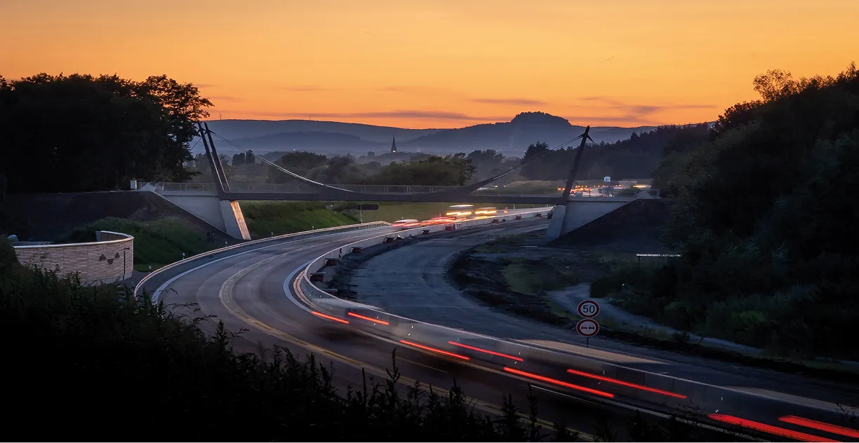 Beleuchtete Autobahn mit einer modernen Hängebrücke bei Sonnenuntergang vor hügeliger Landschaft.