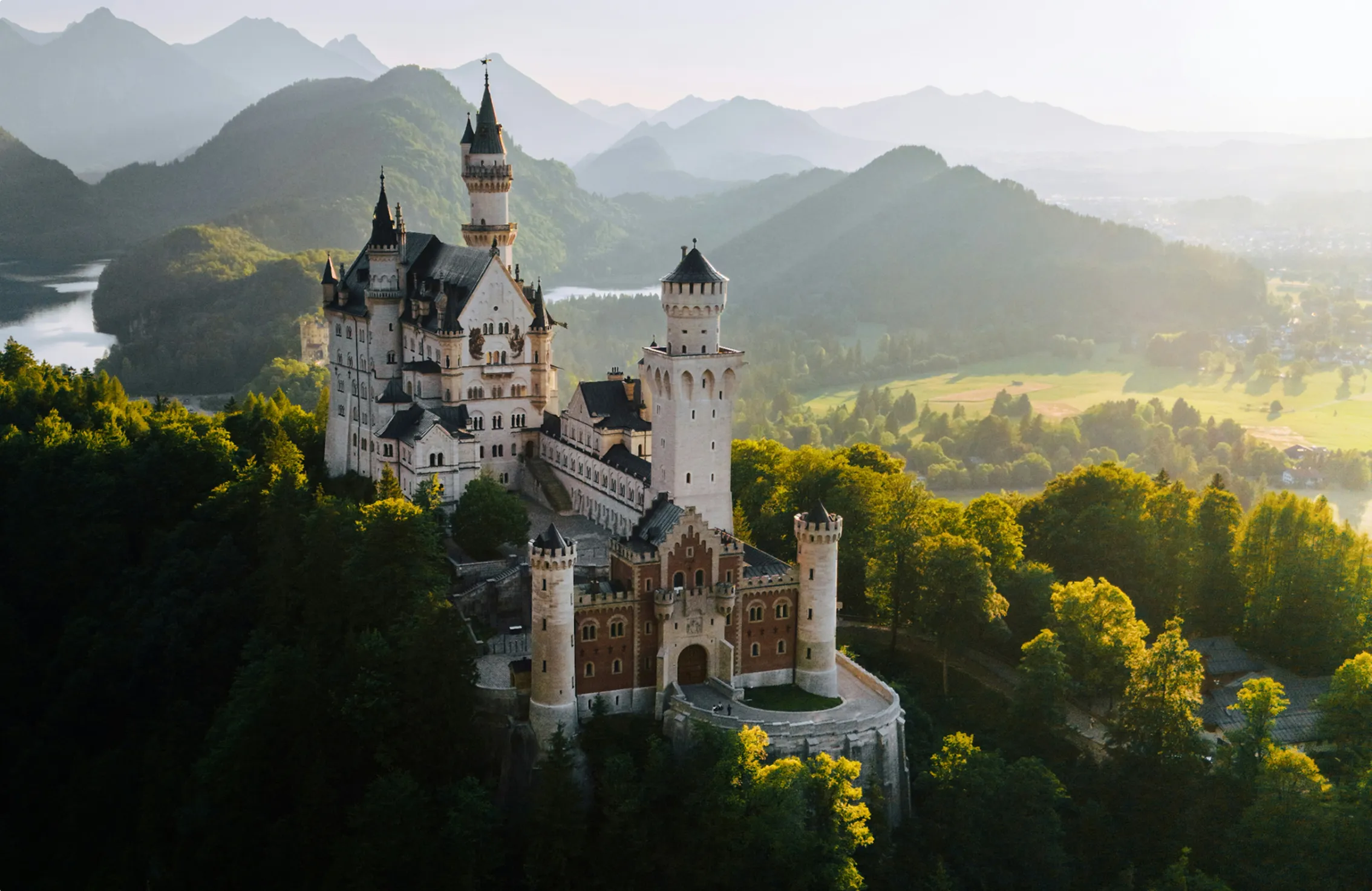 Schloss Neuschwanstein auf einem bewaldeten Hügel mit Bergen und Fluss im Hintergrund im Abendlicht.