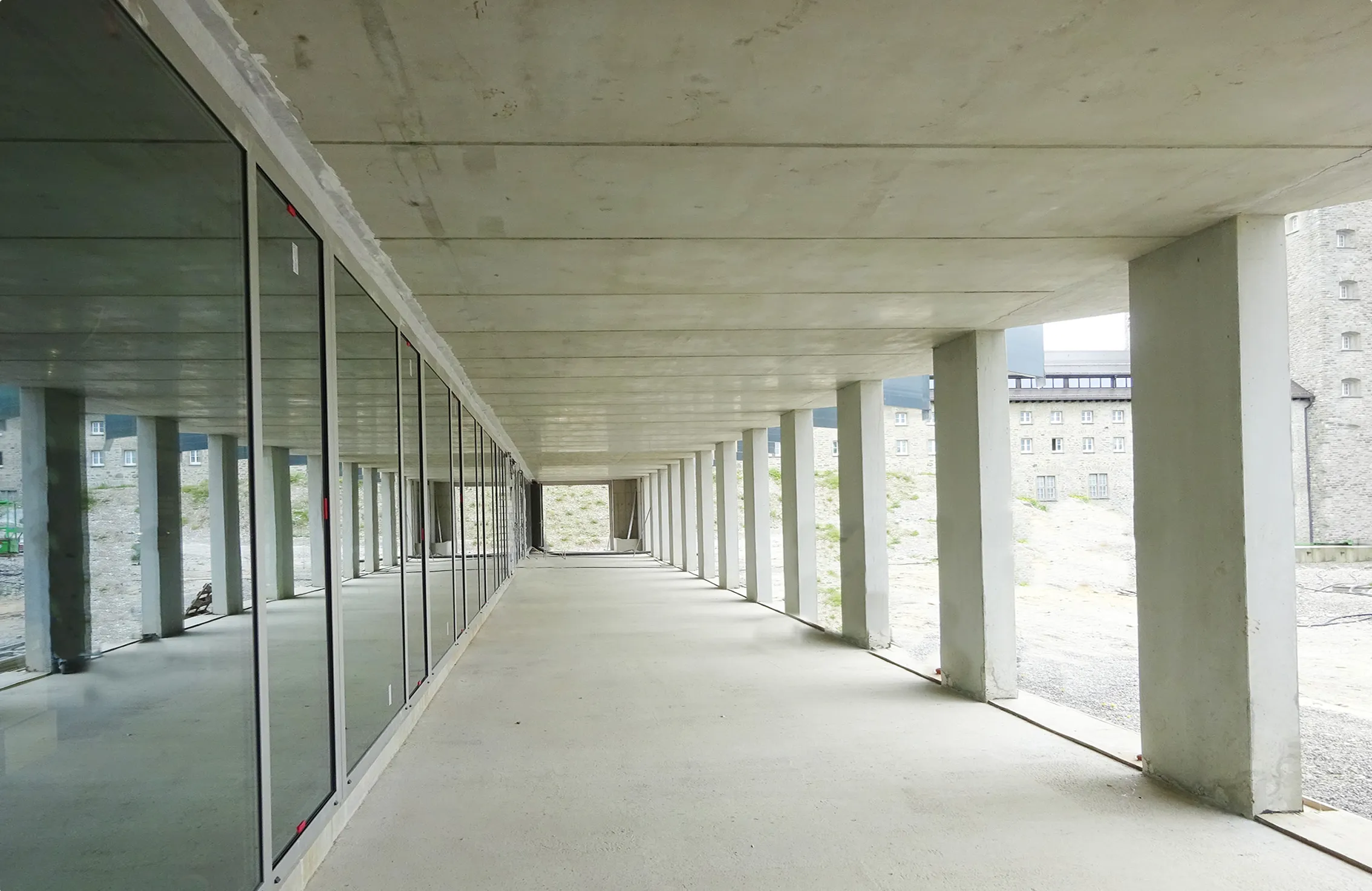 Weitläufiger, leerer überdachter Gang mit Betonboden, rechteckigen Betonsäulen rechts und Glaswänden links, mit Blick auf ein Gebäude im Hintergrund.