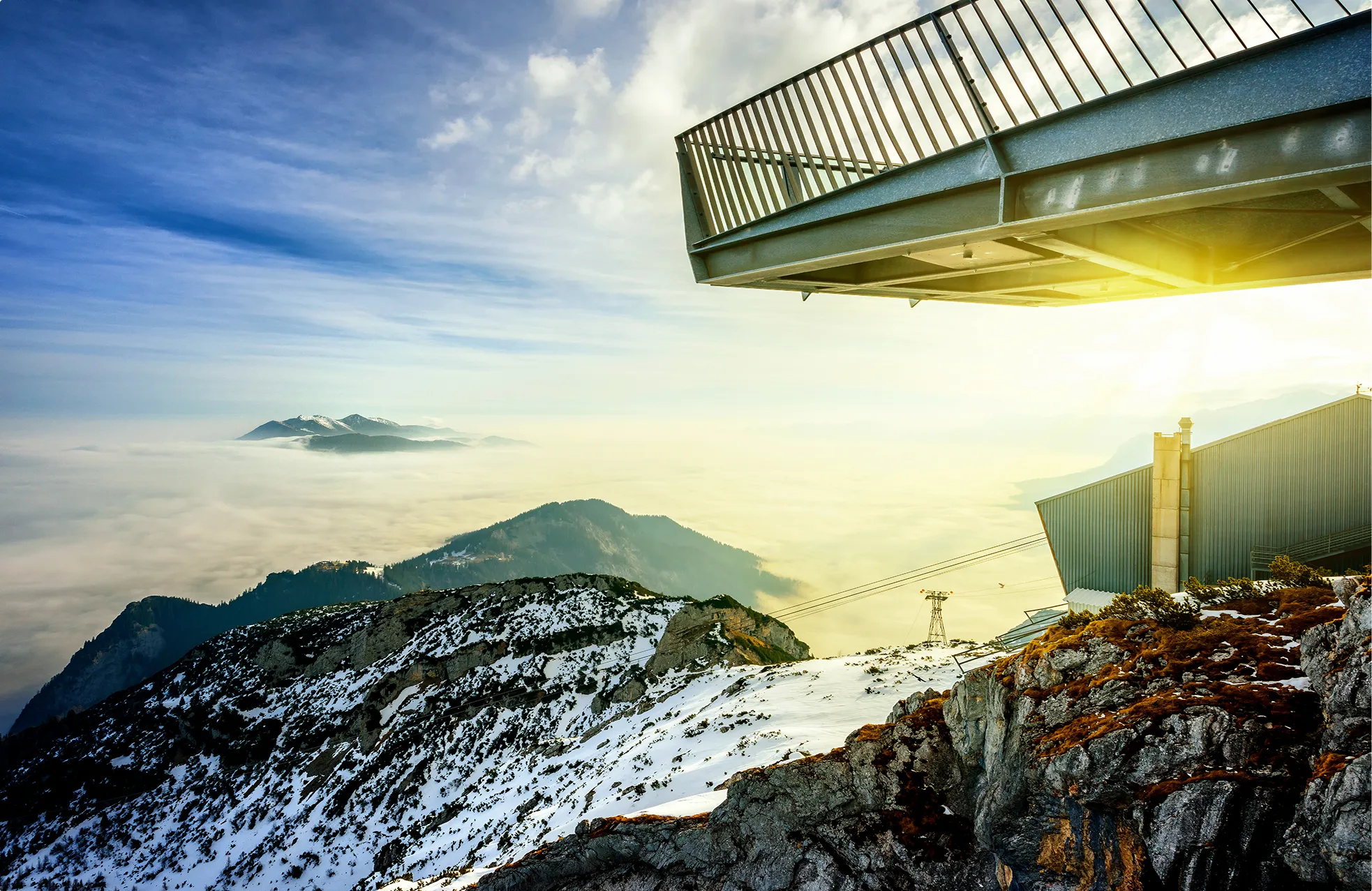 Berggipfel mit Schneeflecken und Felsen bei Sonnenuntergang, darunter liegt ein Wolkenmeer, darüber ragt eine Aussichtsplattform hinaus.