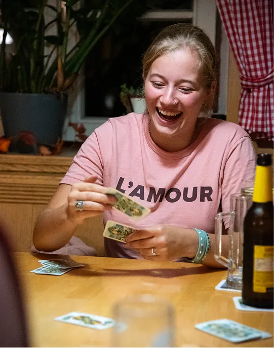 Junge Frau lacht und hält Spielkarten an einem Holztisch mit Getränken.