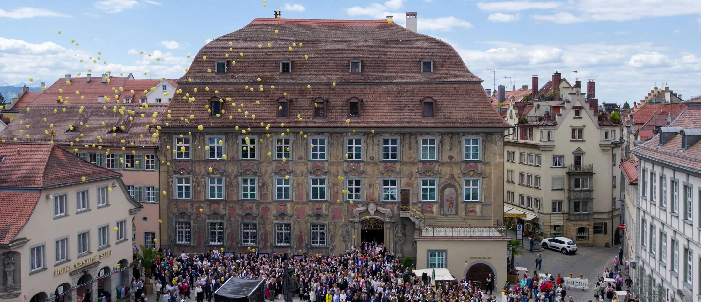 Große Menschenmenge vor einem historischen Gebäude mit verzierten Fassaden, umgeben von alten Häusern mit roten Dächern, während gelbe Luftballons in den Himmel steigen.