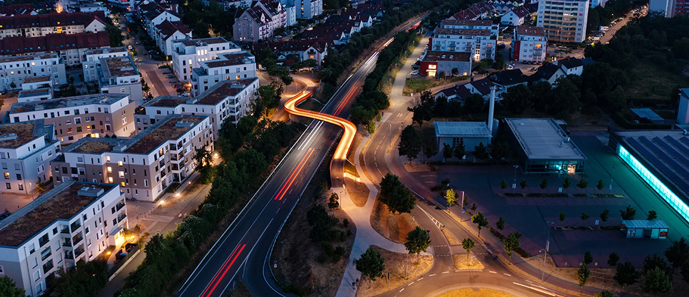 Luftaufnahme einer Stadt bei Nacht mit beleuchteter Fußgängerbrücke über eine Autobahn und Wohngebäuden auf beiden Seiten.