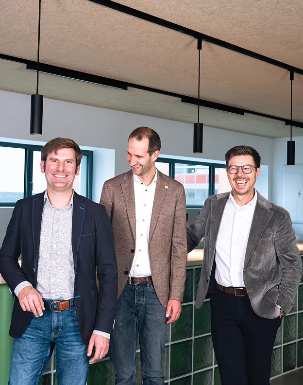 Drei lächelnde Männer in Geschäftskleidung stehen vor einer modernen Büroumgebung mit großen Fenstern und Hängeleuchten.