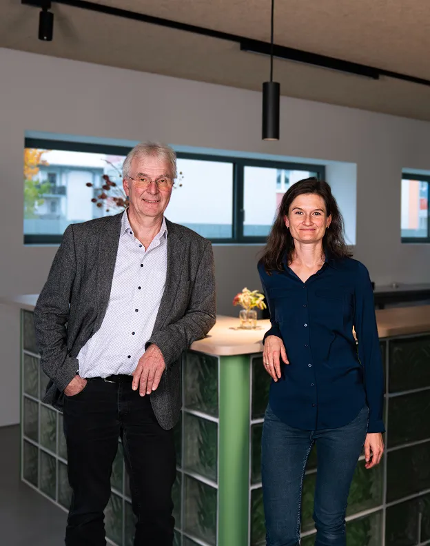 Ein Mann mit grauem Sakko und eine Frau in dunkelblauer Bluse lehnen an einer Tresenfläche aus Glasbausteinen in einem modernen, hellen Raum mit Fenstern.