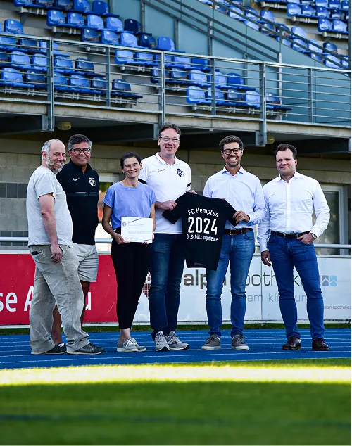 Sechs Erwachsene stehen auf einer Laufbahn im Stadion, eine Person hält ein Schild, eine andere zeigt ein Trikot mit der Aufschrift 'FC Kempten 2024 Dr.Schütz Ingenieure'.