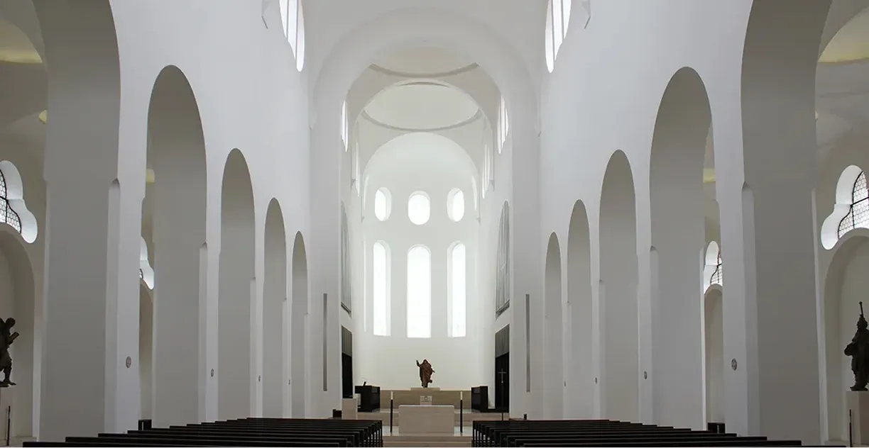 Innenansicht einer weißen Kirche mit hohen Rundbogenfenstern und Statuen am Altar.