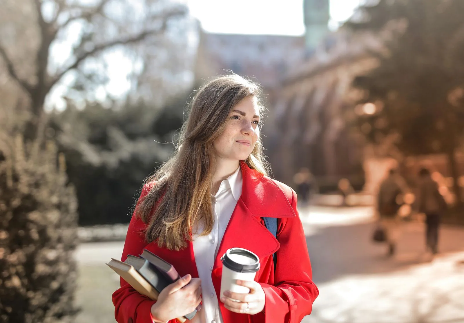 A schoolgirl stands outdoors holding books in one hand and a coffee cup in the other