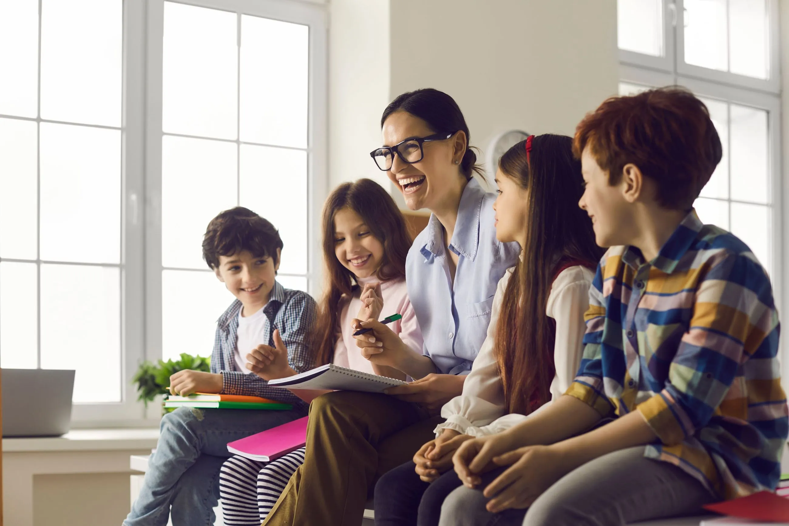 Nauczycielka siedząca z dziećmi przy oknie w klasie, uśmiechnięta podczas wspólnej rozmowy.