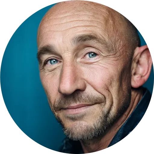 Portrait rapproché d’un homme chauve aux yeux bleus et barbe légère, sourire discret, fond bleu.