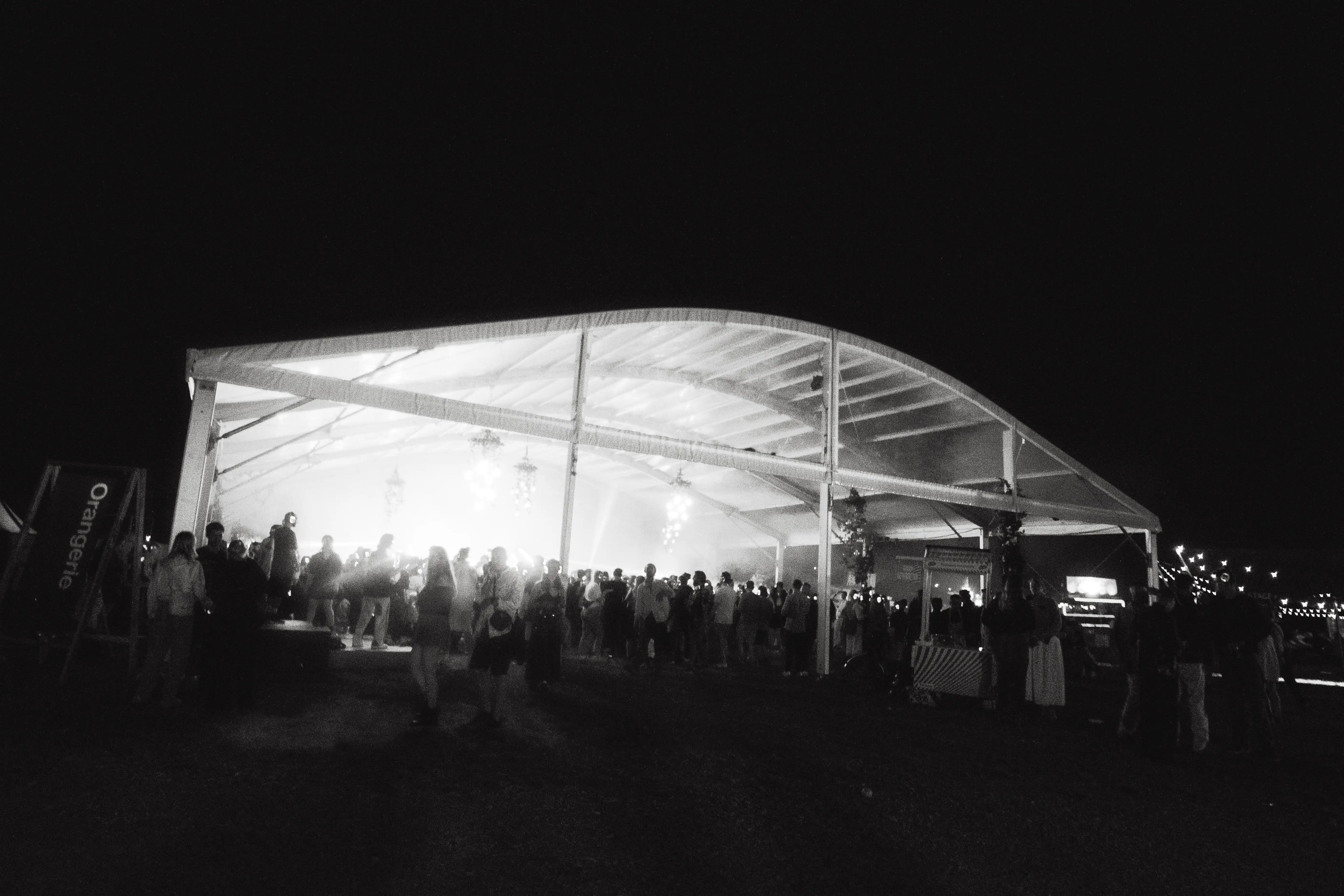 Orangerie stage seen from afar, at night. Bright lights come from its core. People stand near it and all around.
