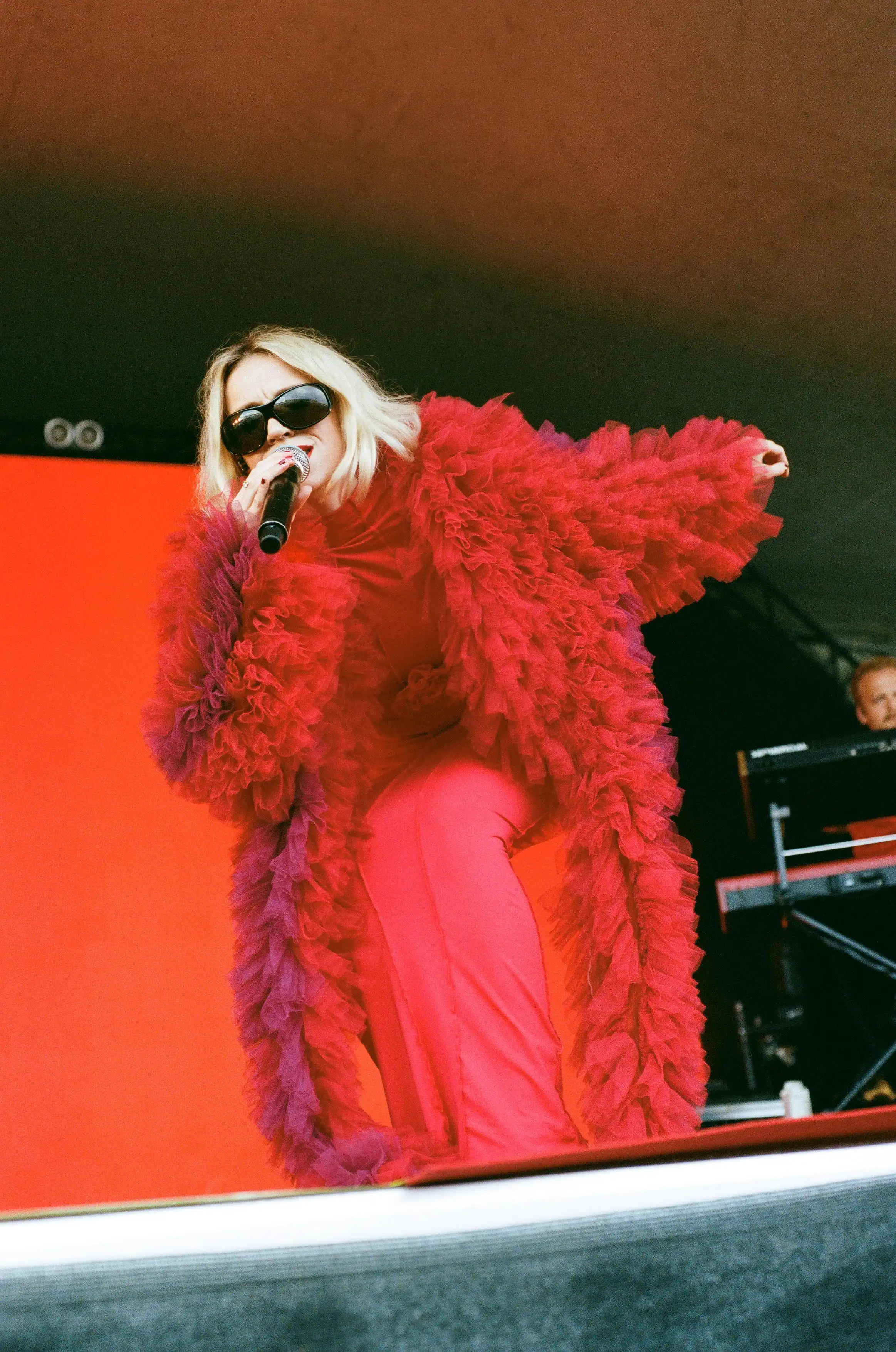 Rmma Sehested Høeg performing at O Days, wearing a long, fluffy red jacket, a long red body-con dress and big, black sunglasses.