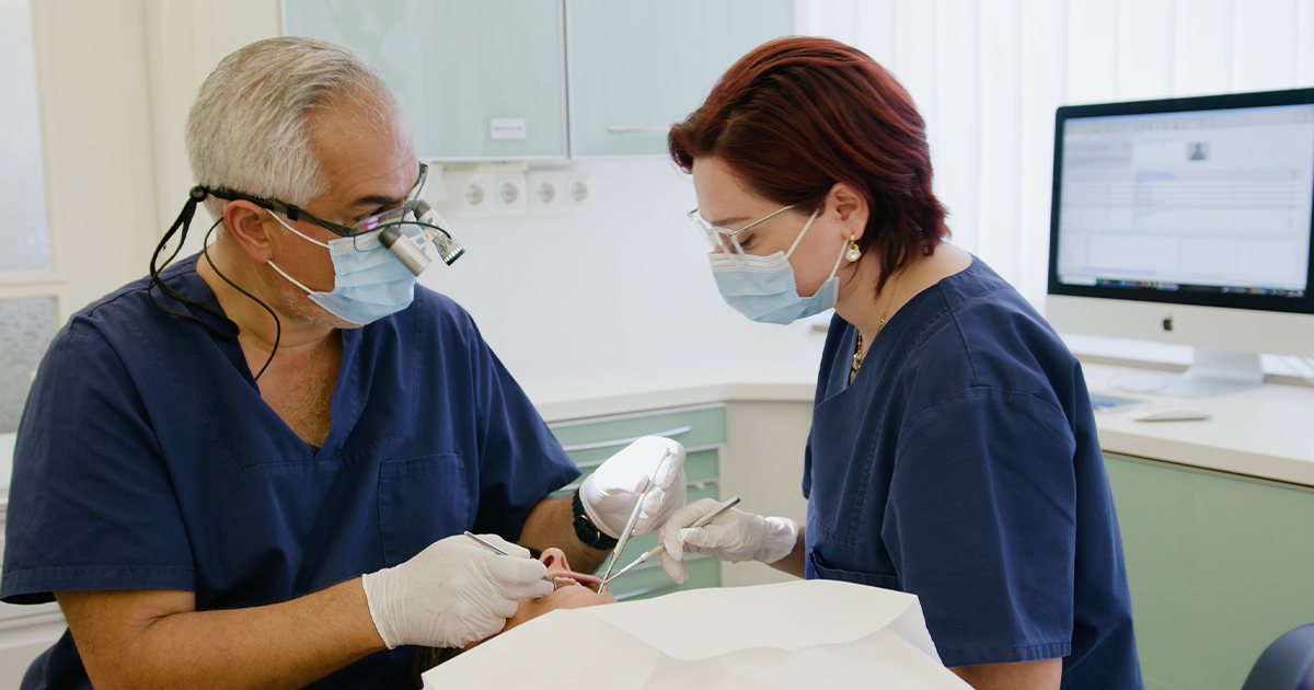 Das Socrates Dental Team. Der Zahnarzt Sivan Ates und acht Mitarbeiter in blauen Arbeitskleidung posieren in einer hellen Zahnarztpraxis um einen Zahnarztstuhl.