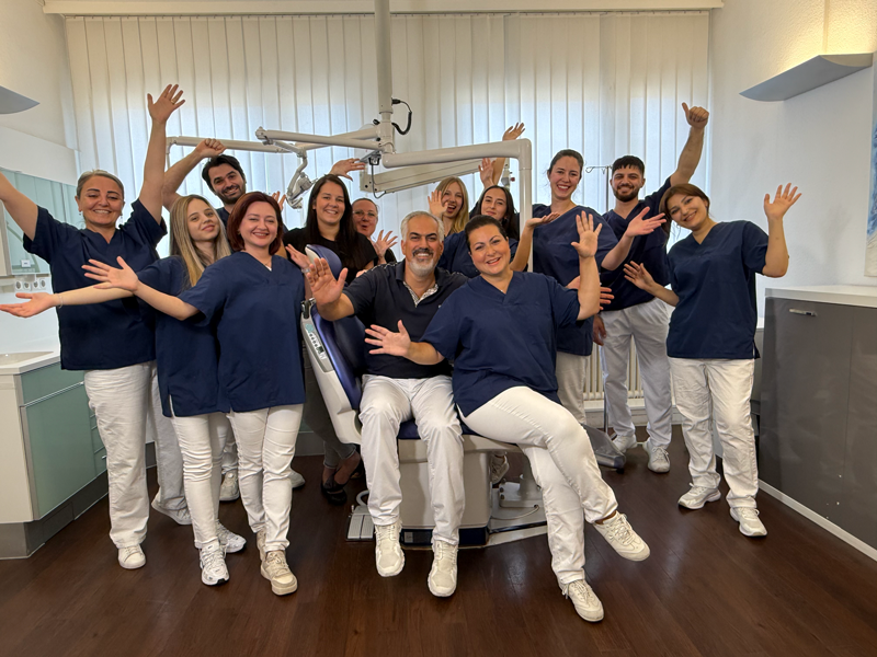 Das Socrates Dental Team. Der Zahnarzt Sivan Ates und acht Mitarbeiter in blauen Arbeitskleidung posieren in einer hellen Zahnarztpraxis um einen Zahnarztstuhl.