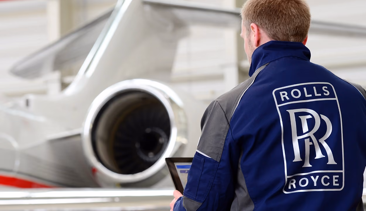 Luft- und Raumfahrtingenieur mit dem Rücken zur Kamera, das Rolls-Royce-Logo auf seinem Overall und ein Flugzeug im Hintergrund