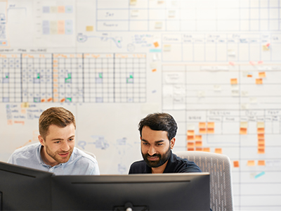 Two colleagues looking at computer screens with a wall full of data behind them