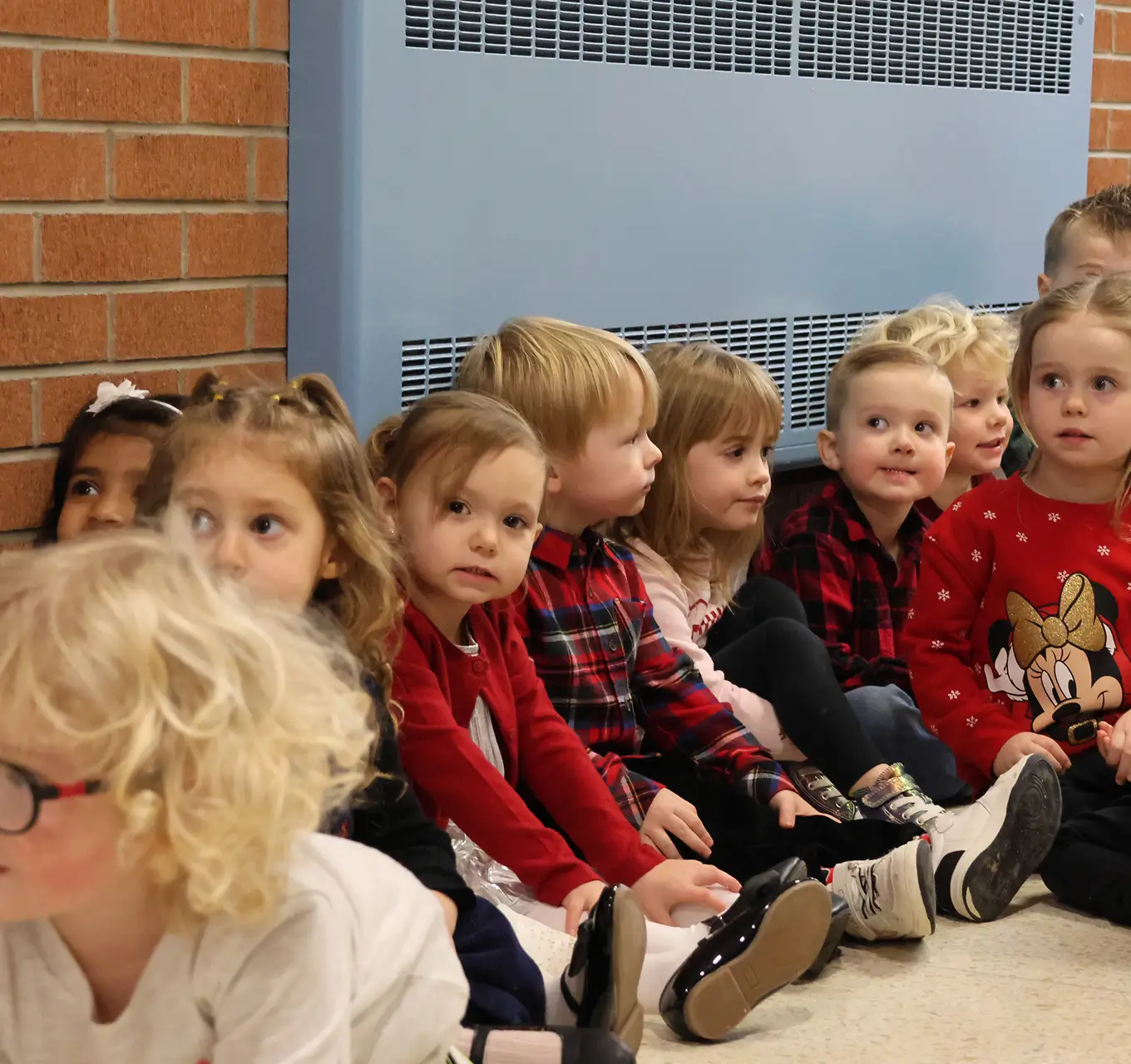 A group of young children in colorful clothing sitting on the floor indoors, looking attentive.