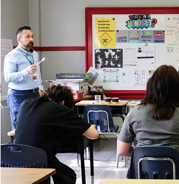 A teacher stands at the front of a classroom, holding a spoon and speaking to students.
