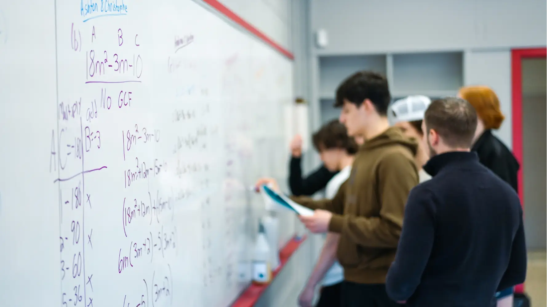 Five students solve math problems on a large whiteboard in a classroom.