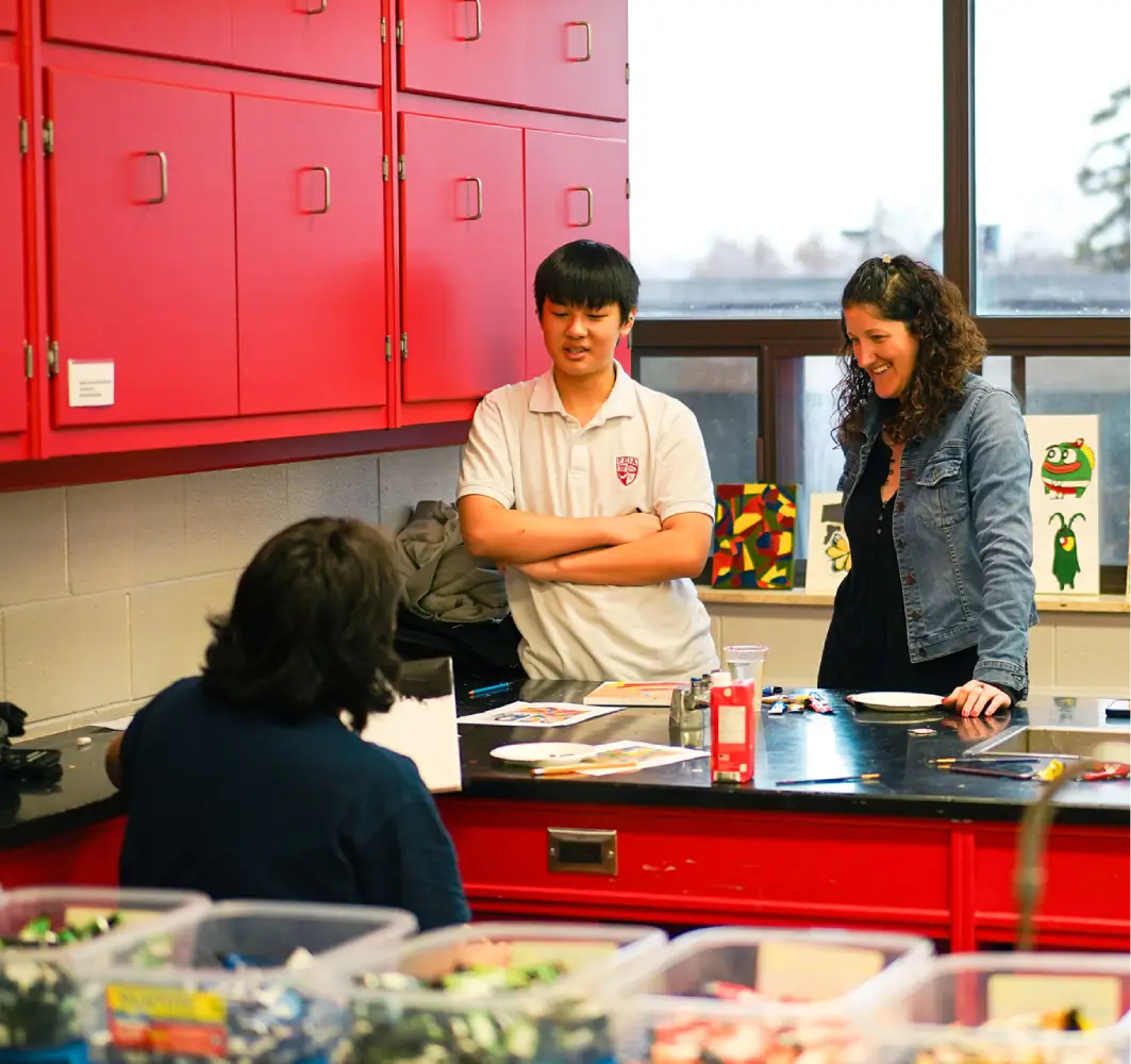 Two people stand talking to someone seated at a desk in a brightly lit classroom.