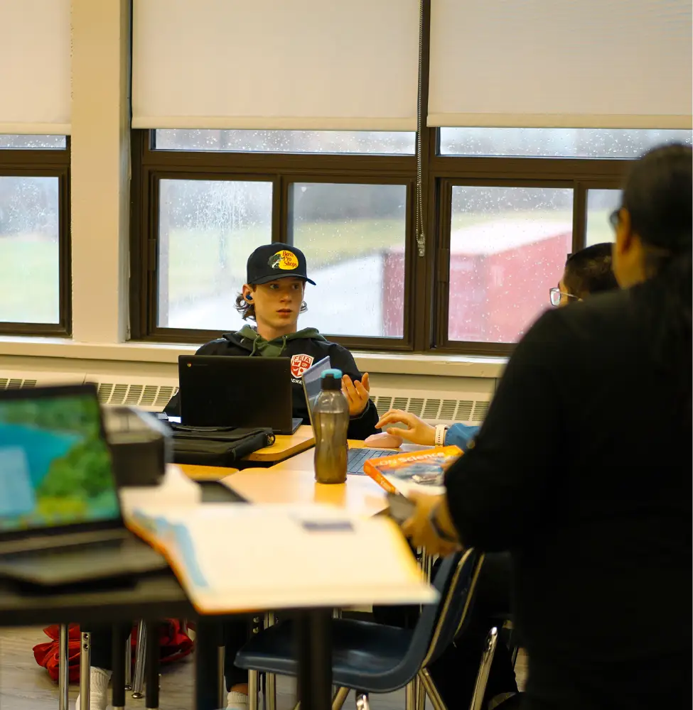 A student wearing a cap speaks while sitting at a classroom table with laptops and books.