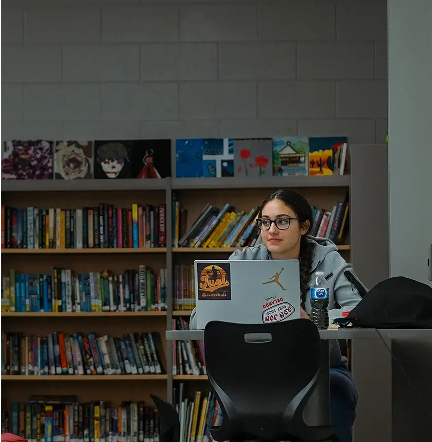A person sits at a desk in a library, using a laptop covered with stickers.