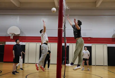Two people jump to block a volleyball over a net in a gym setting.