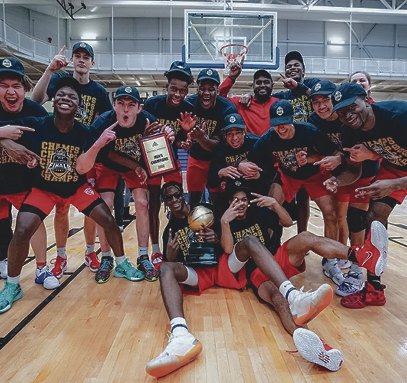 A jubilant basketball team celebrates, holding a trophy and plaque, wearing championship shirts on a court.