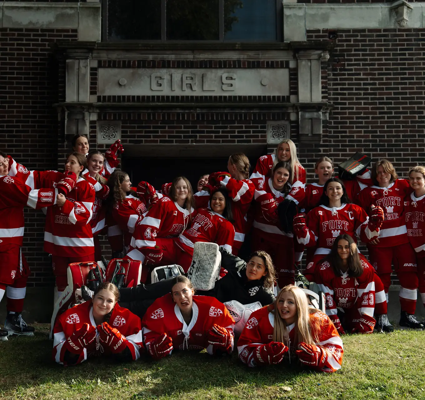 A girls' hockey team in red uniforms poses happily in front of a building.