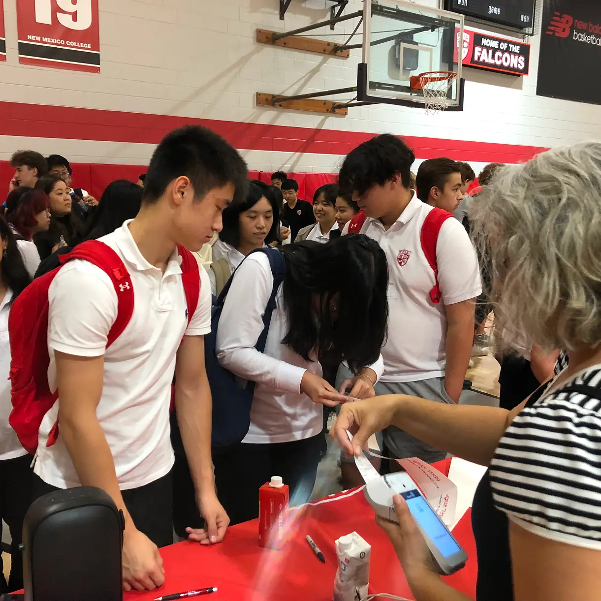 A group of students in uniforms gather at a table, interacting with a person handing out papers.