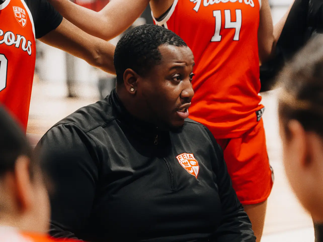 A coach talks to basketball players in orange jerseys during a timeout.