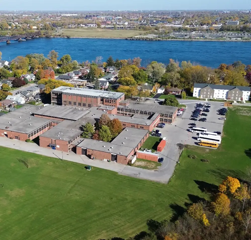 Aerial view of a large brick school building surrounded by trees and a river in the background.