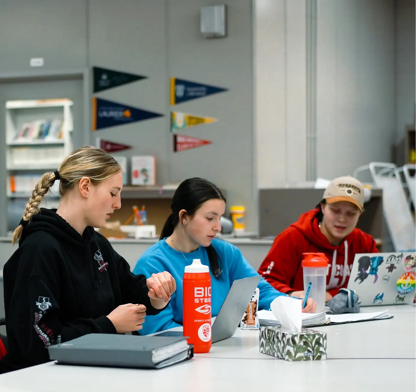 Three people sitting at a table, working on laptops and taking notes in a classroom setting.