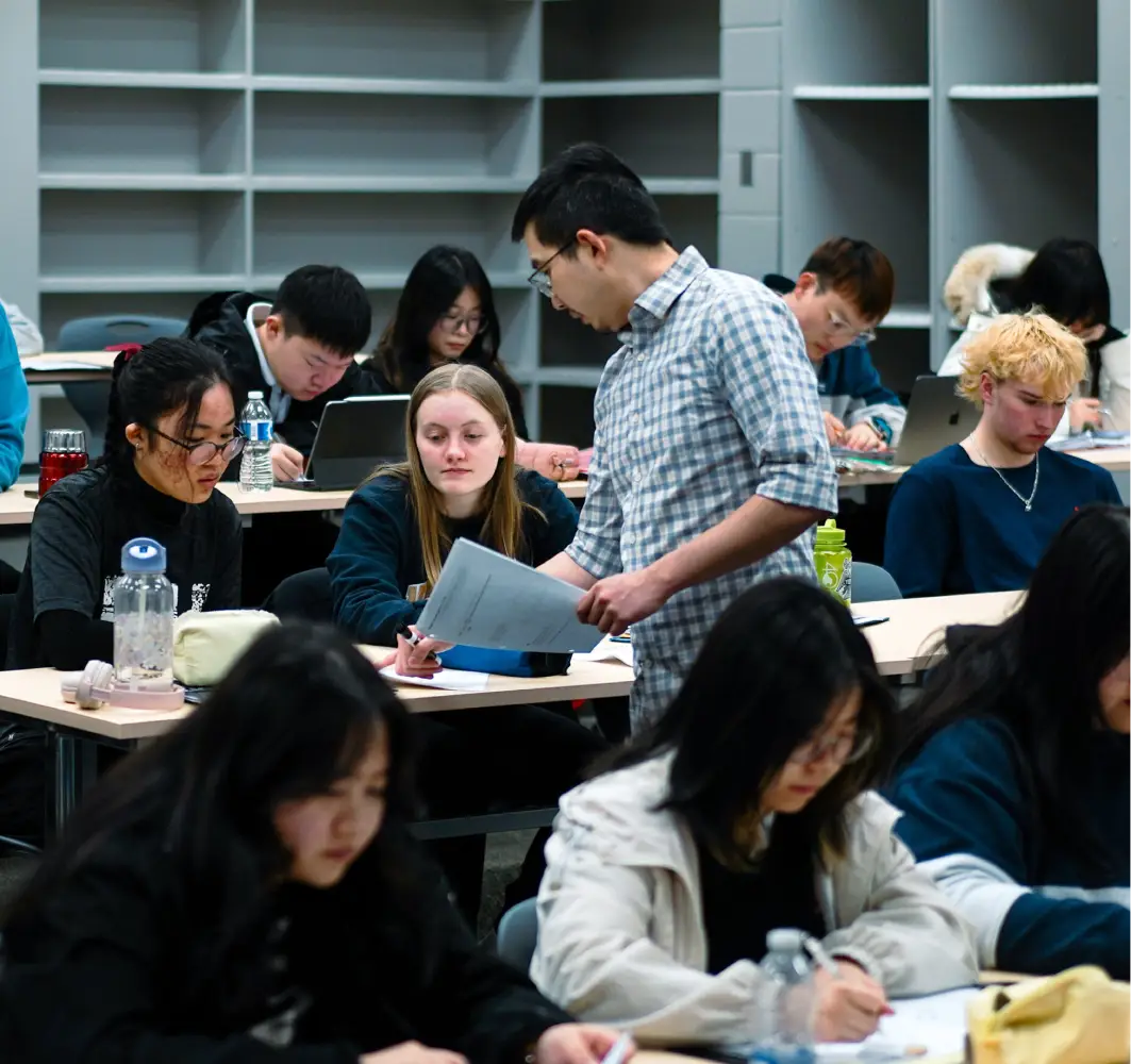 A teacher assists a student with classwork in a busy classroom with other students working.