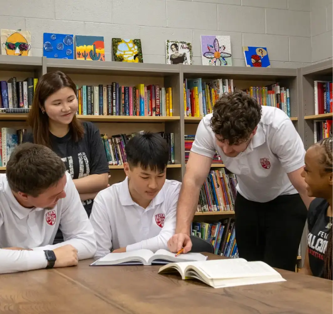 A group of students in white uniforms discusses a book at a library table.