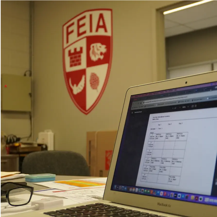 Open laptop on a desk with a schedule, near a wall with a FEIA logo.