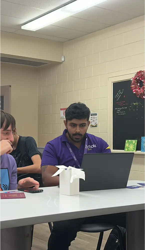 Man focused on laptop, sitting at a table with others in a casual setting.