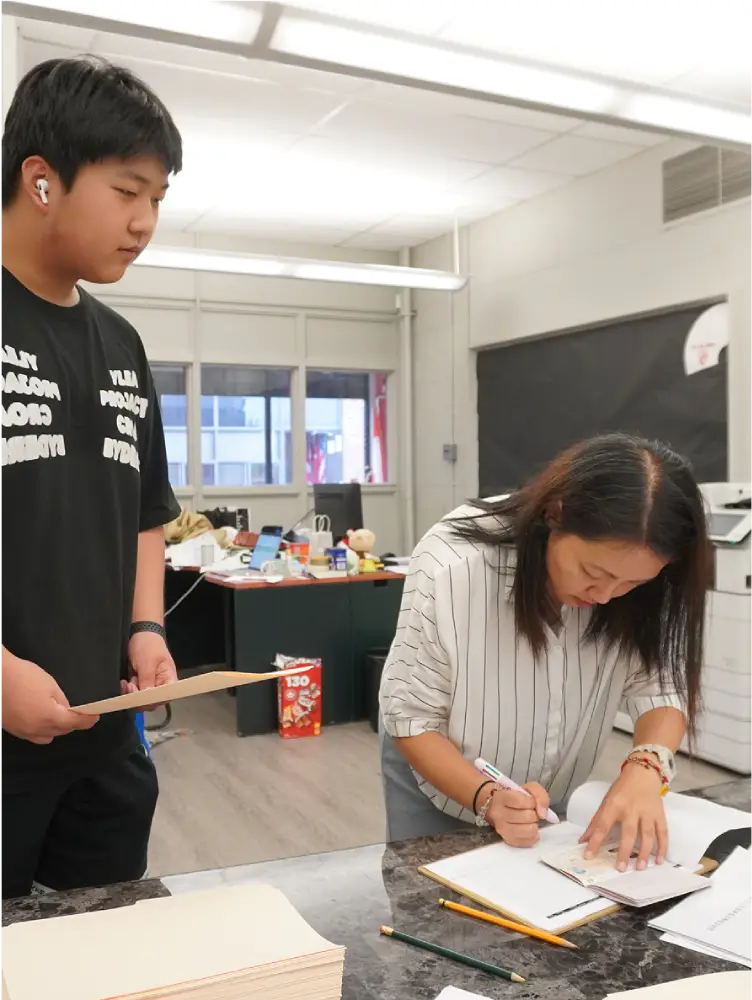 A woman is sitting at a desk writing, while a boy stands nearby holding papers.