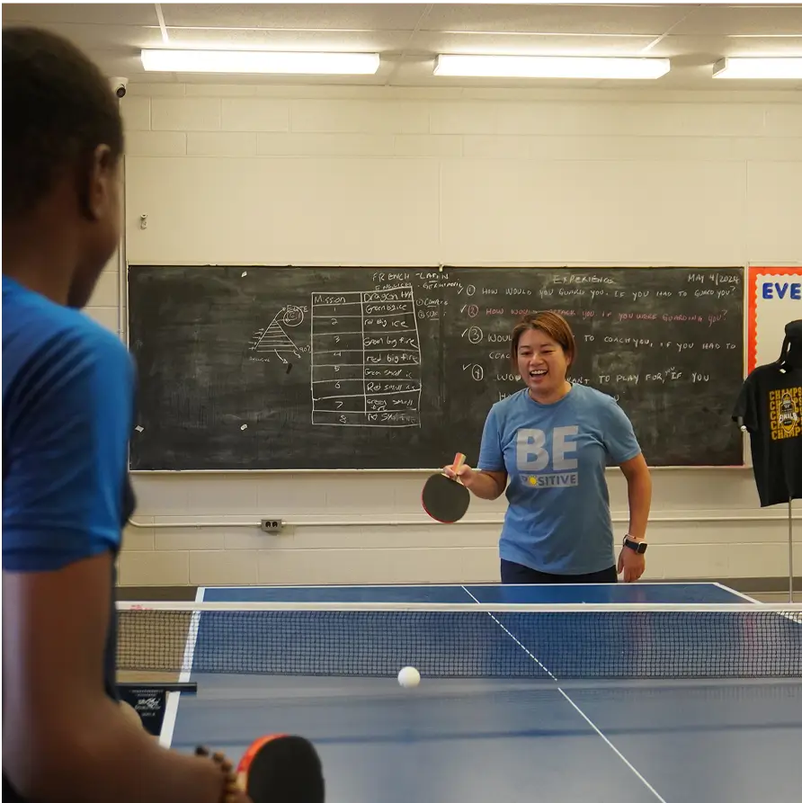 Two people playing table tennis in a classroom with a chalkboard in the background.