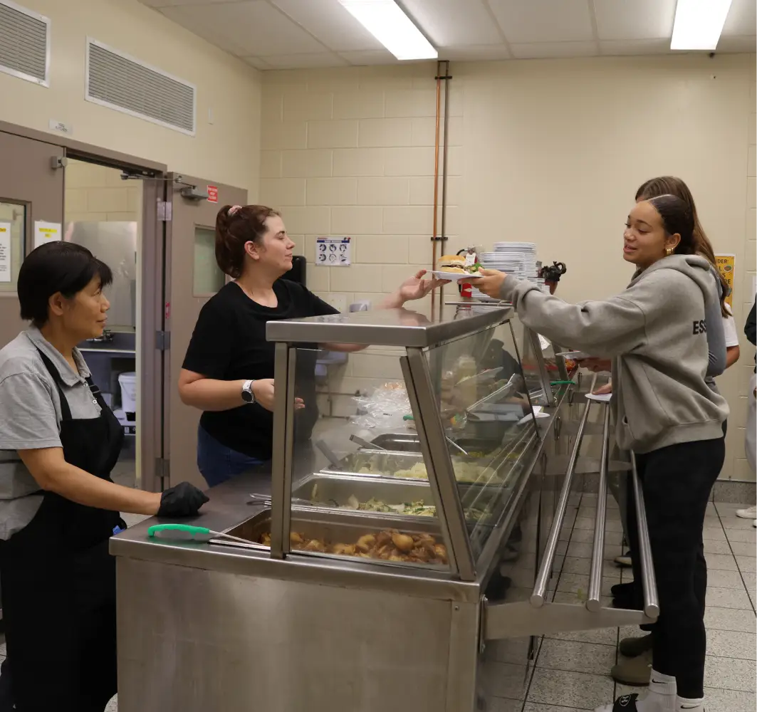 Two cafeteria workers serve food to a smiling student holding a plate at the counter.