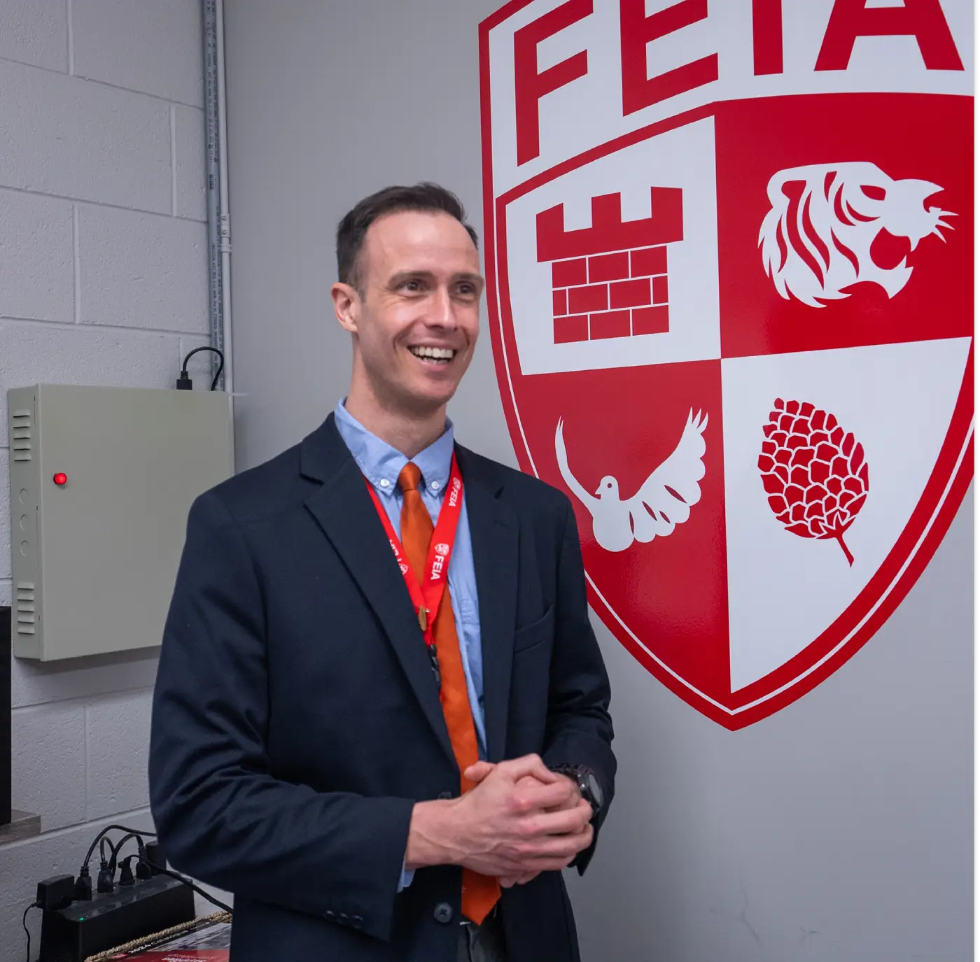 Man in a suit smiles while standing next to a red and white emblem on a wall.