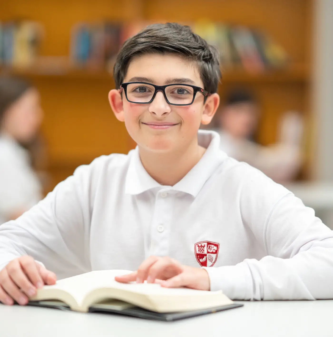 A young student in glasses reads a book at a library table.