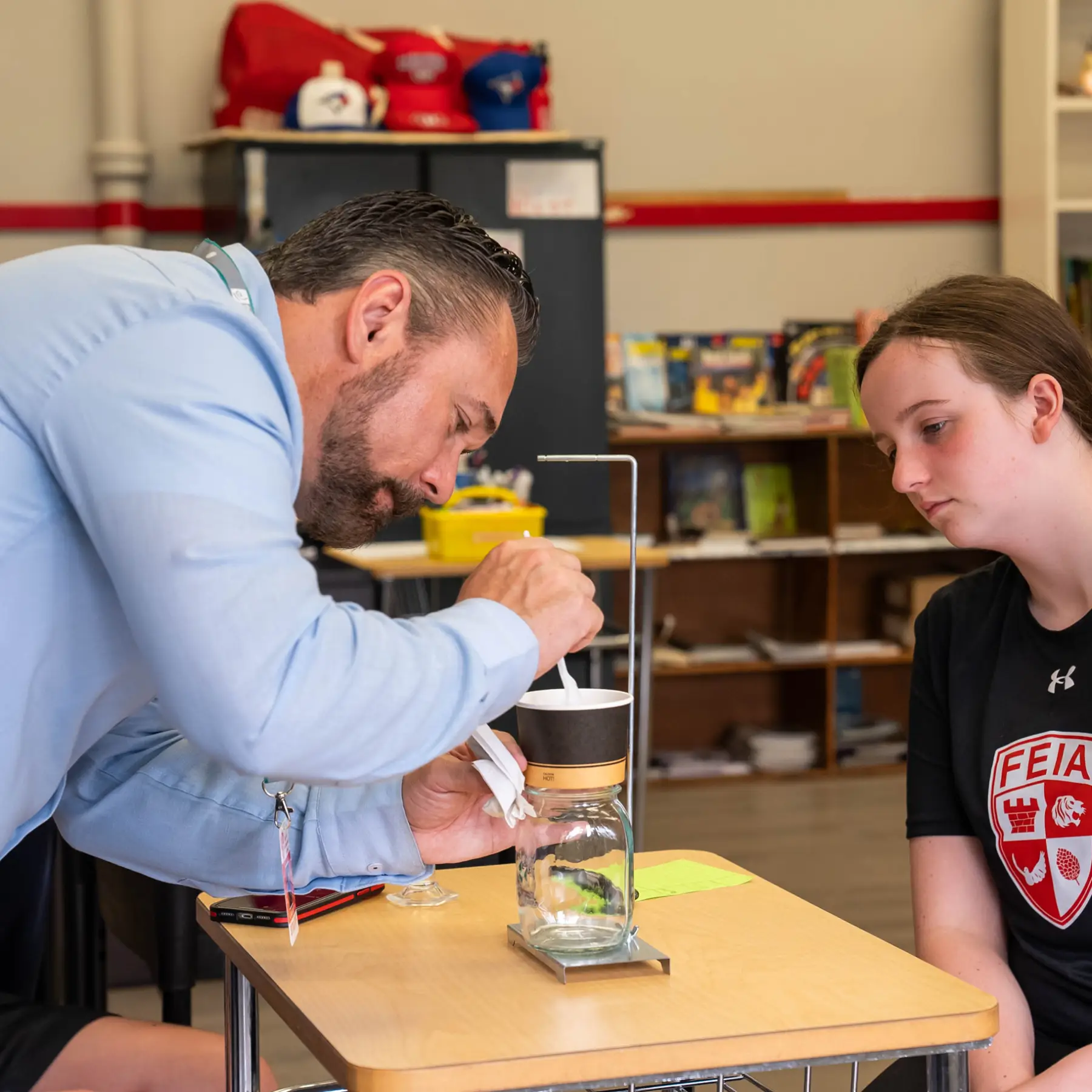 A teacher demonstrates an experiment to a student using a jar and a small apparatus on a desk.