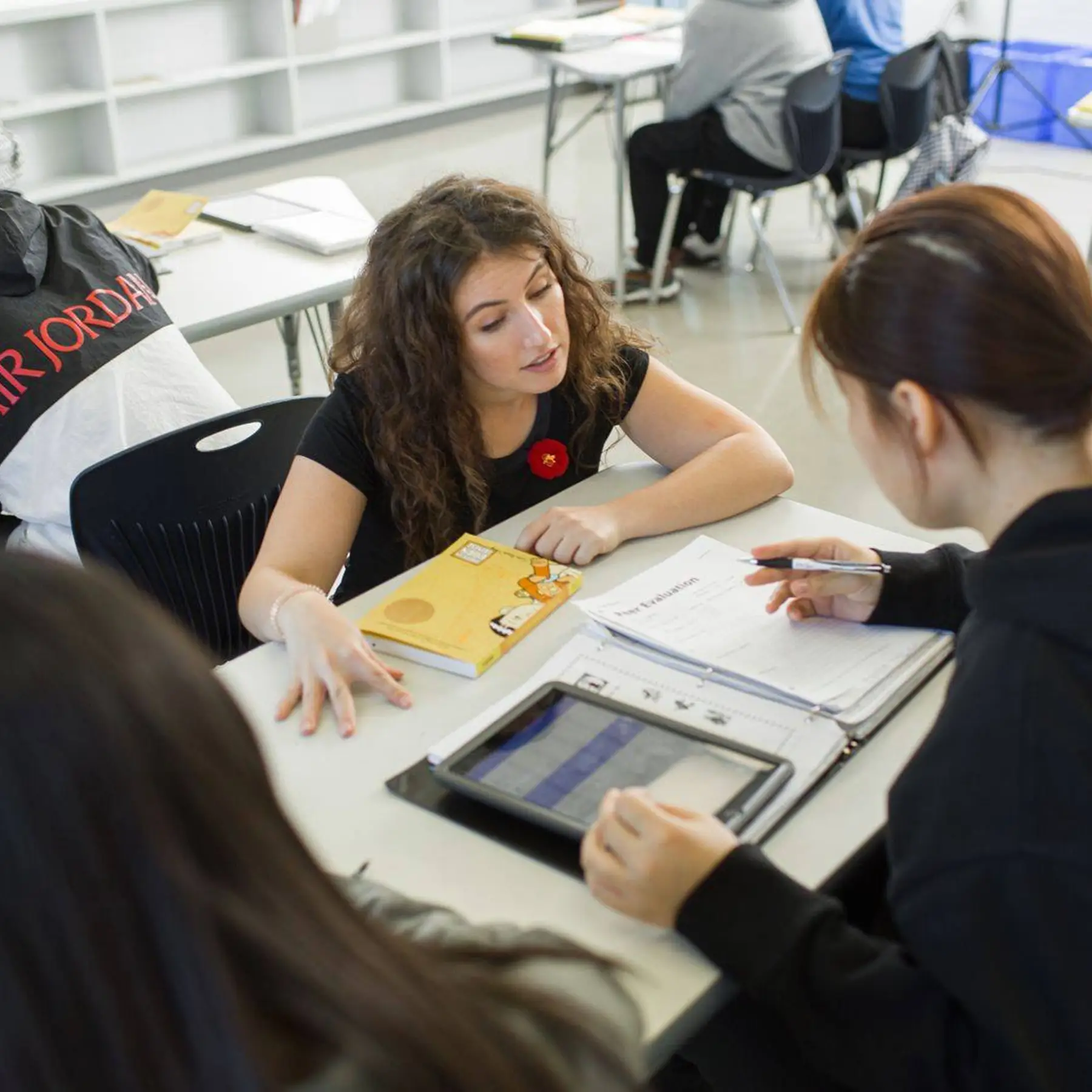 A woman with curly hair speaks to a group seated at a table in a classroom.