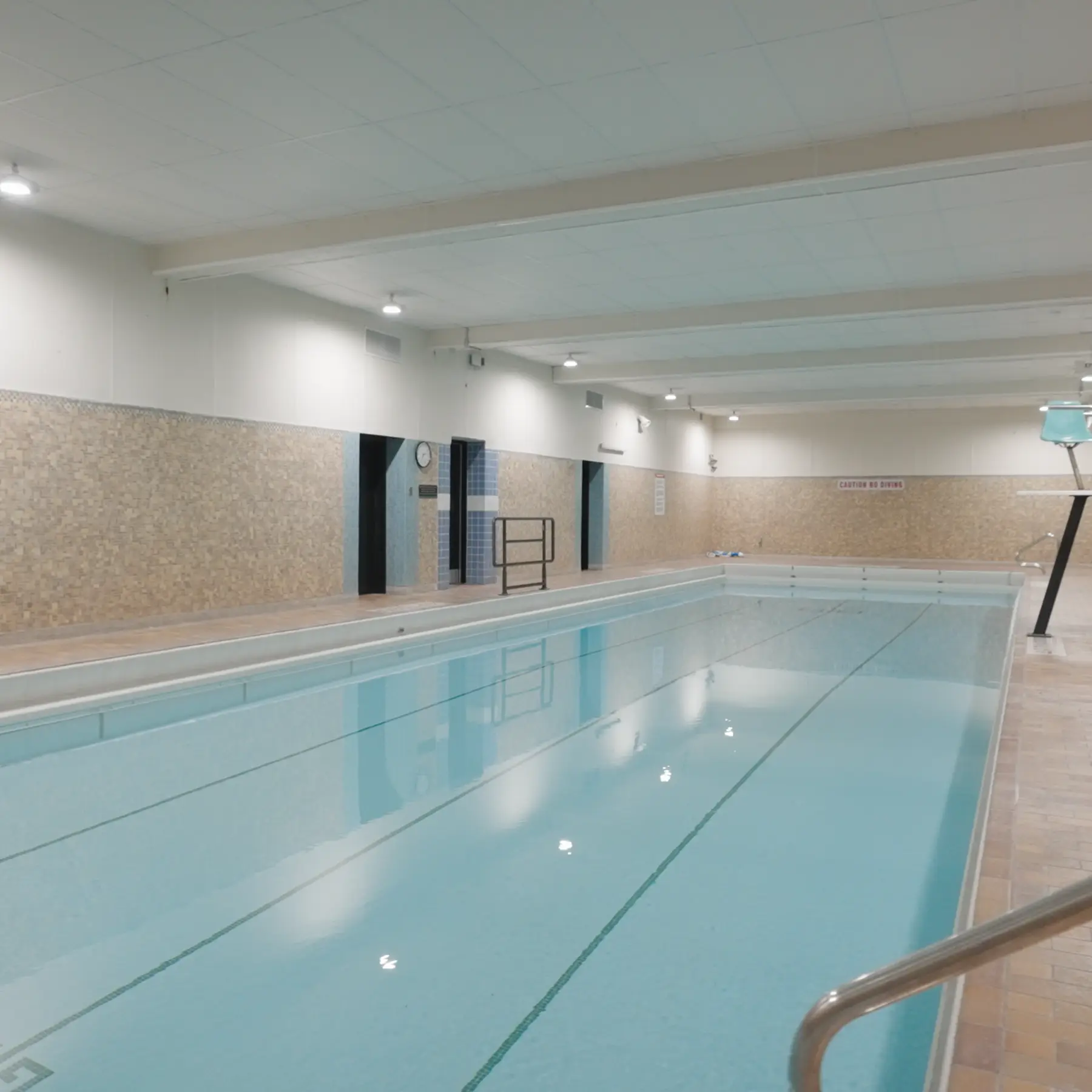 Indoor swimming pool with clear water, surrounded by tiled walls and ceiling lights.