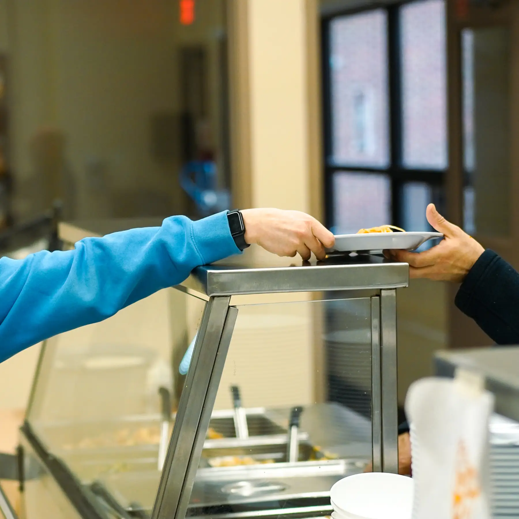 A person in a blue sweater hands a tray with food to another person across a counter.